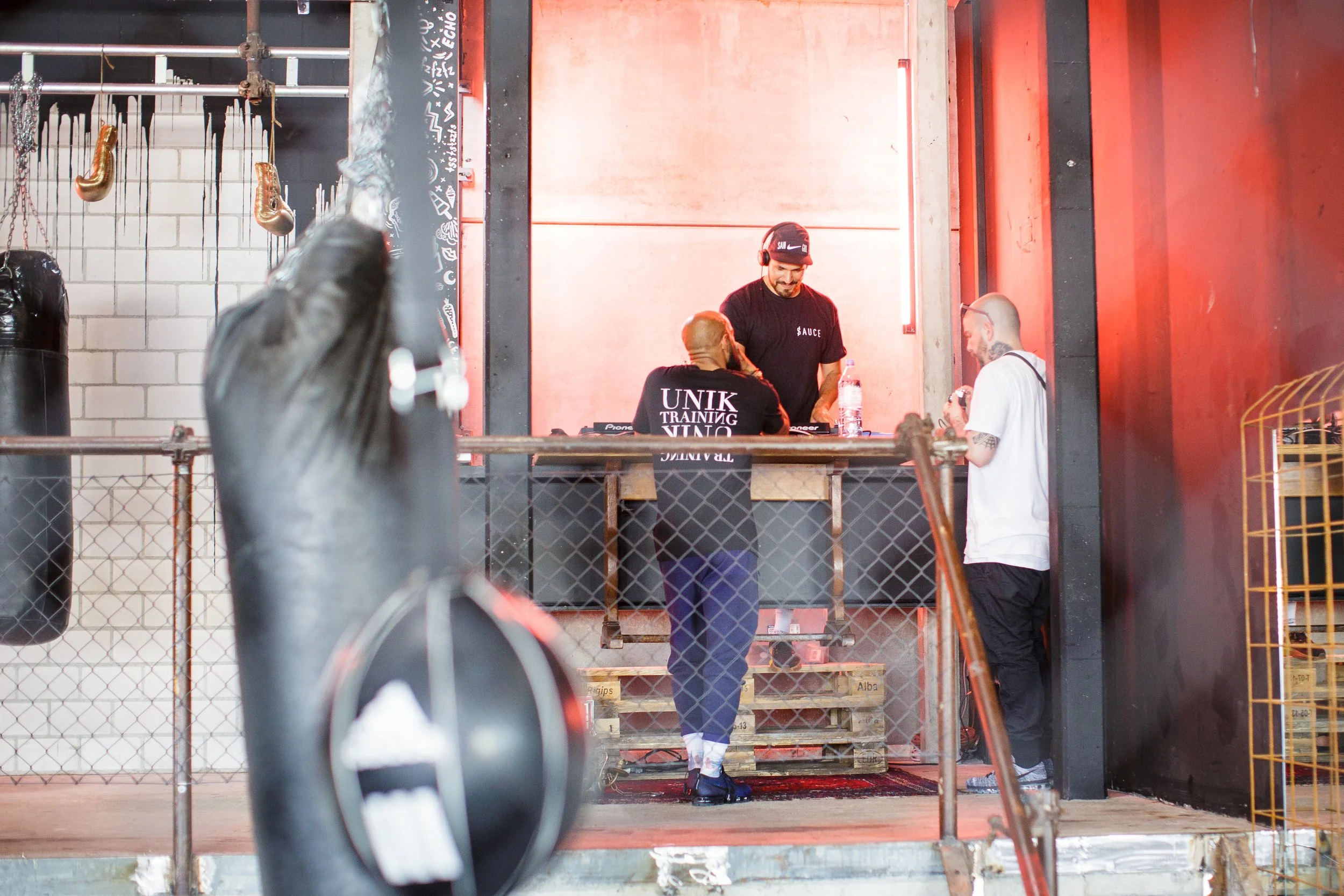 Zwei Männer mit DJ-Equipment hinter einer Bar, ein Mann mit T-Shirt, das 'UNIK TRAINING 'VINO' trägt, und ein Mann mit Brille und Tattoo, sprechen mit einem DJ, im Hintergrund rote Wände in einem Fitnessstudio oder Club.