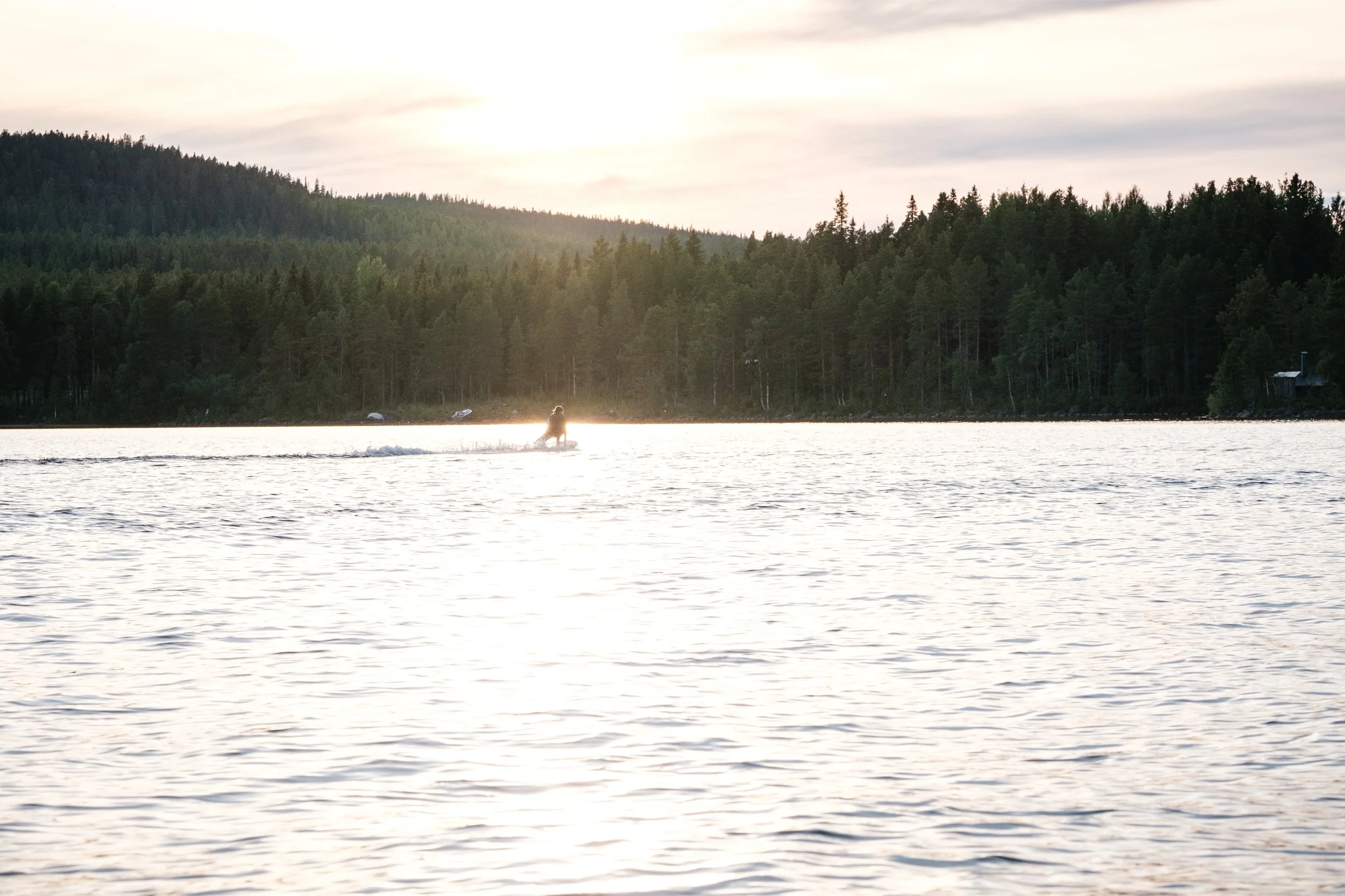Ein Person fährt mit einem Boot auf einemsee bei Sonnenuntergang, im Hintergrund sind Bäume und eine bewölkte Himmel.