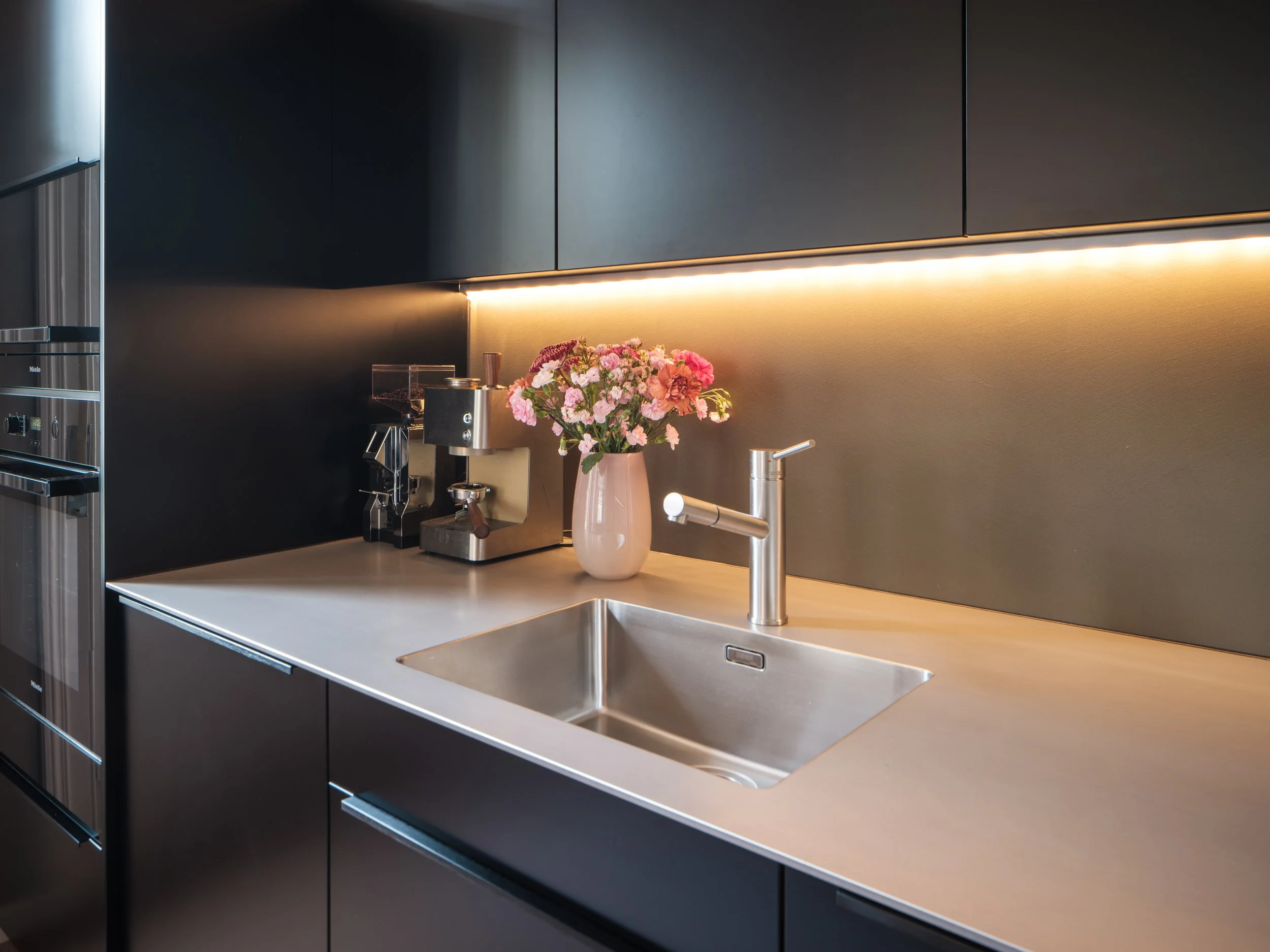 Moderne Küchenspüle mit blumen, Wasserhahn, Kaffeeautomaten und schwarzen Schränken, mit warmem Licht.