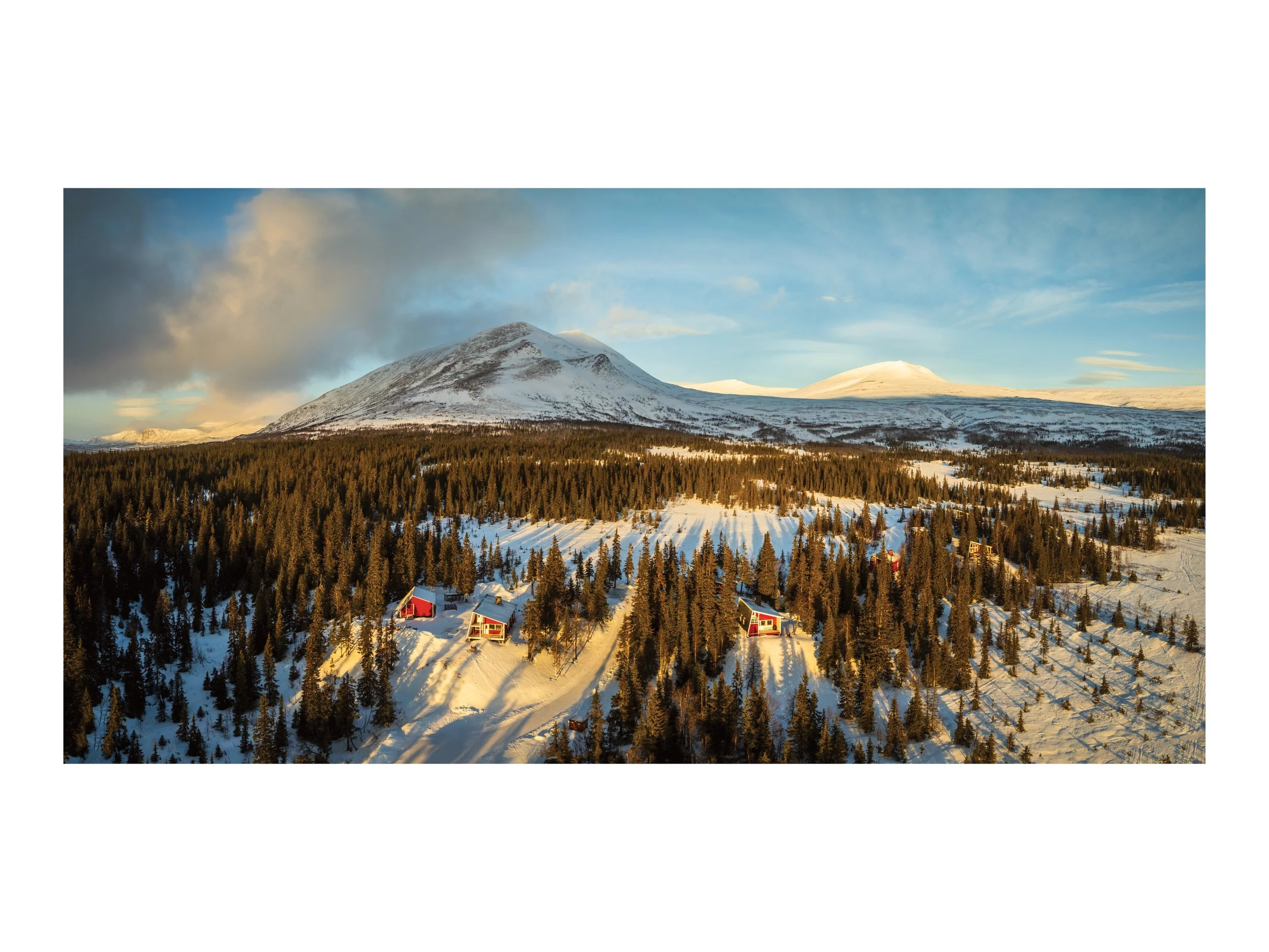 Vogelperspektive auf verschneite Berge und einen Wald mit roten Häusern in Norwegen bei Sonnenuntergang.