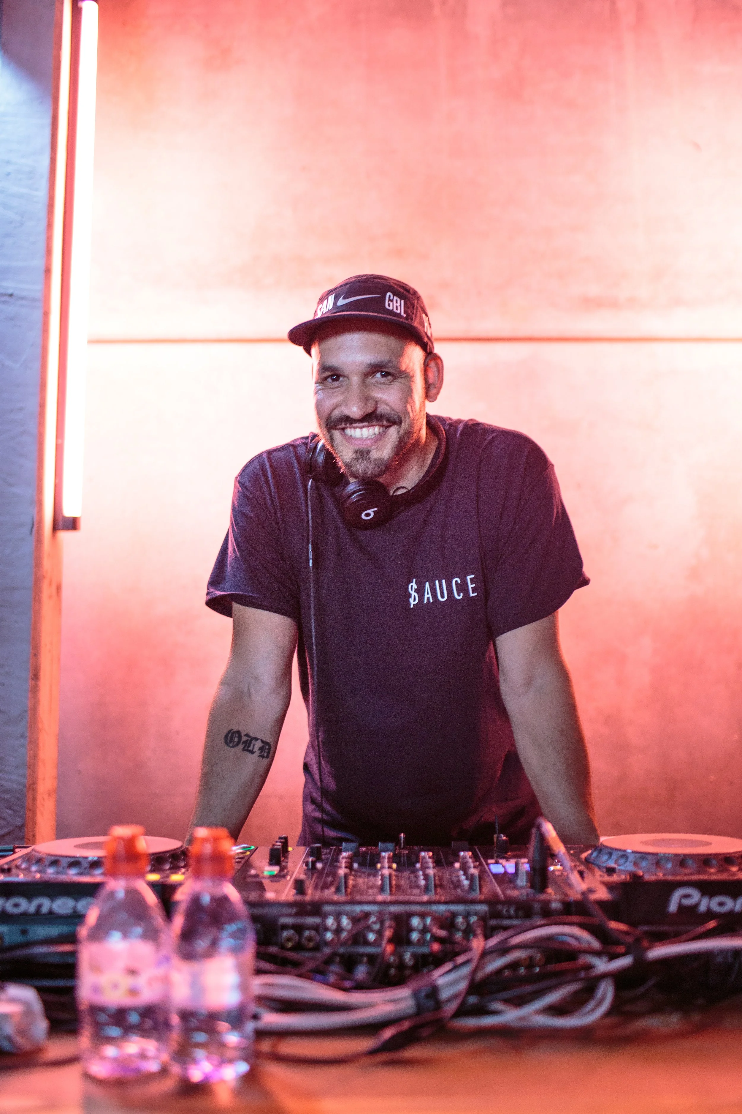 Ein DJ mit Kopfhörern steht hinter einem DJ-Setup bei einer Party, mit Wasserflaschen im Vordergrund und einer warmen Beleuchtung im Hintergrund.