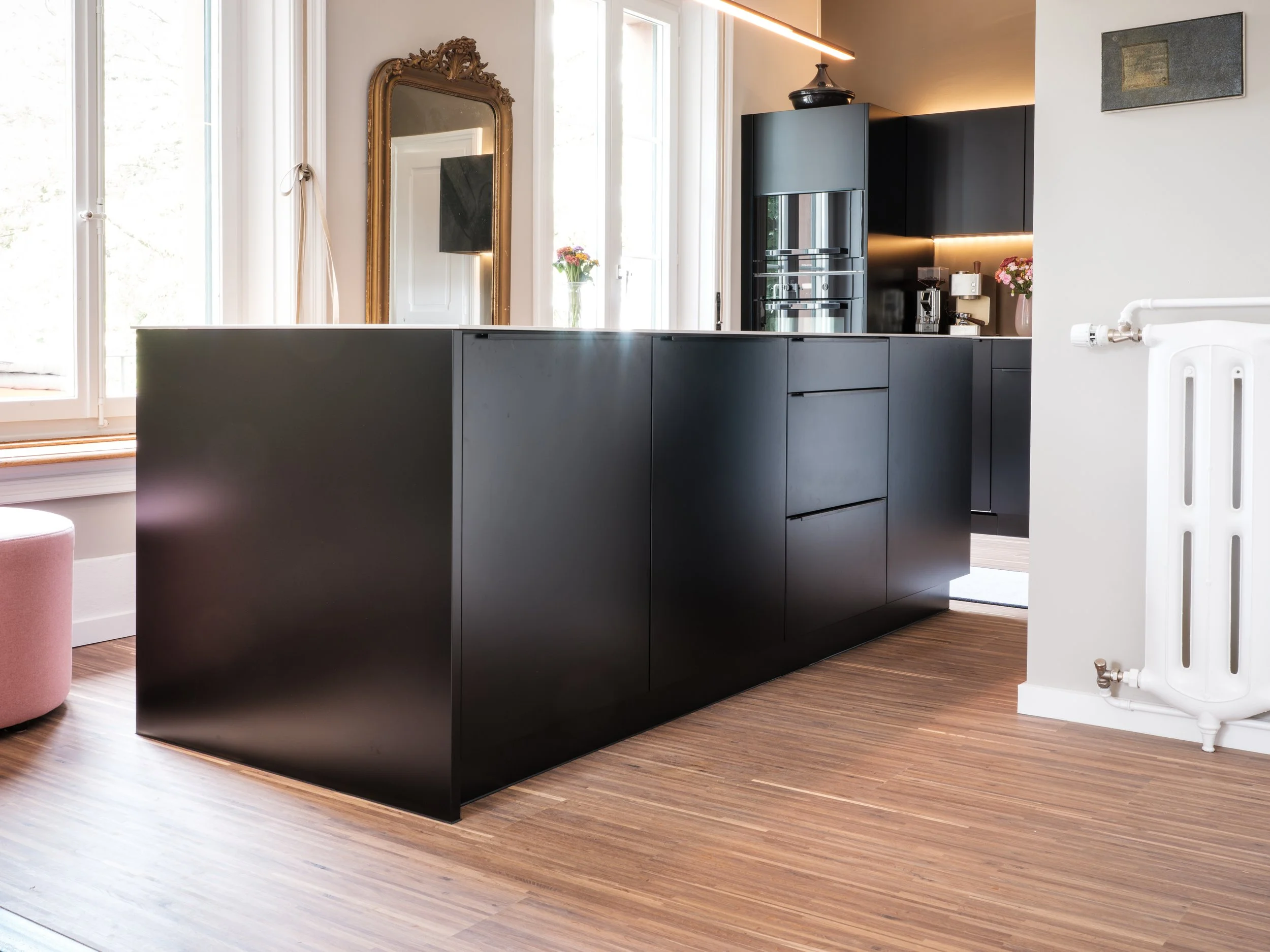 Moderne schwarze Kücheninsel mit Schubladen und Stauraum, Hintergrund mit schwarzen Küchenschränken, holzfarbener Boden, große Fenster, Spiegel im Vintage-Stil, Blumen, Kaffeemaschine, Wasserkocher.