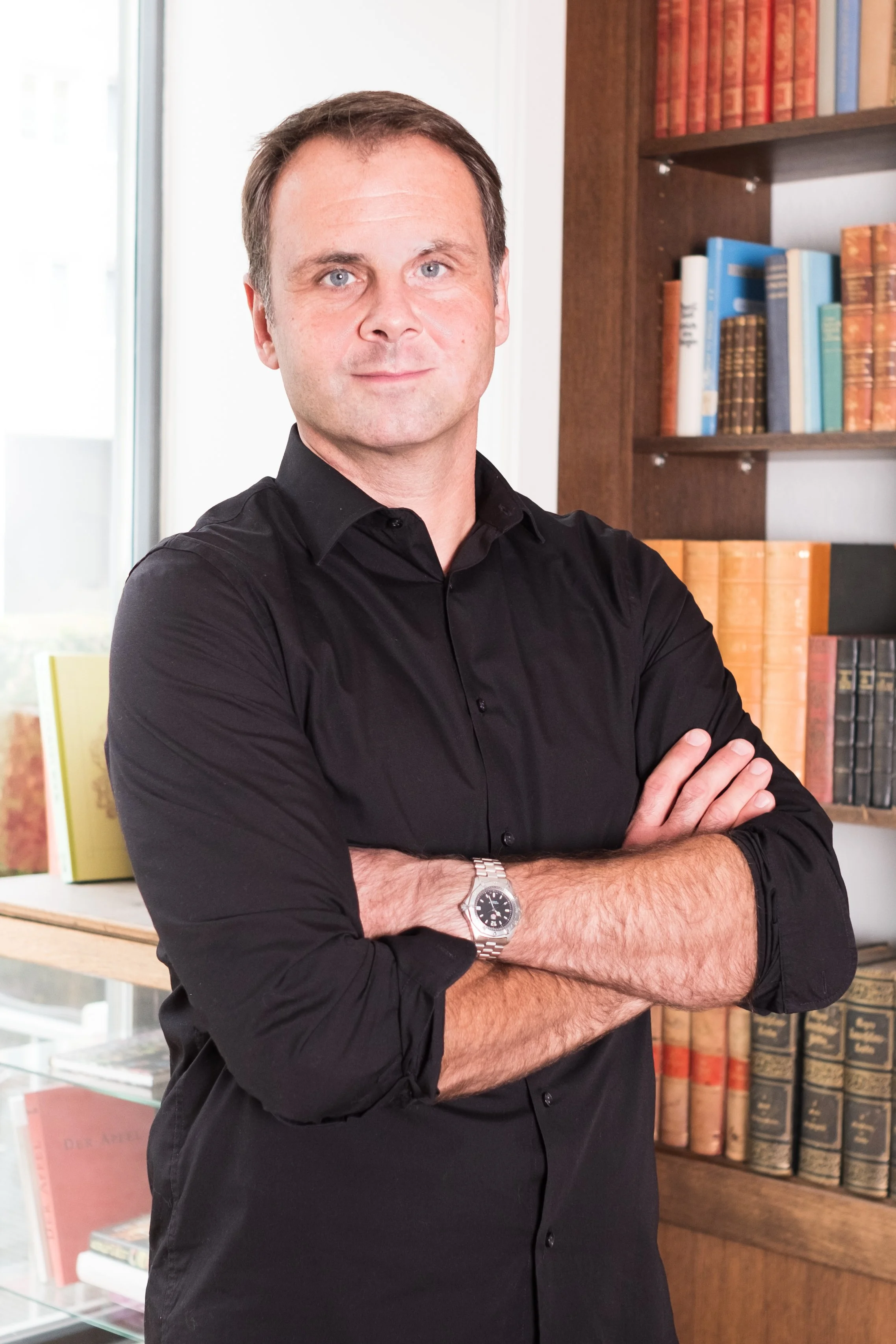 Ein Mann mit schwarzen Hemd, silberner Armbanduhr, steht vor Bücherregal in Raum.