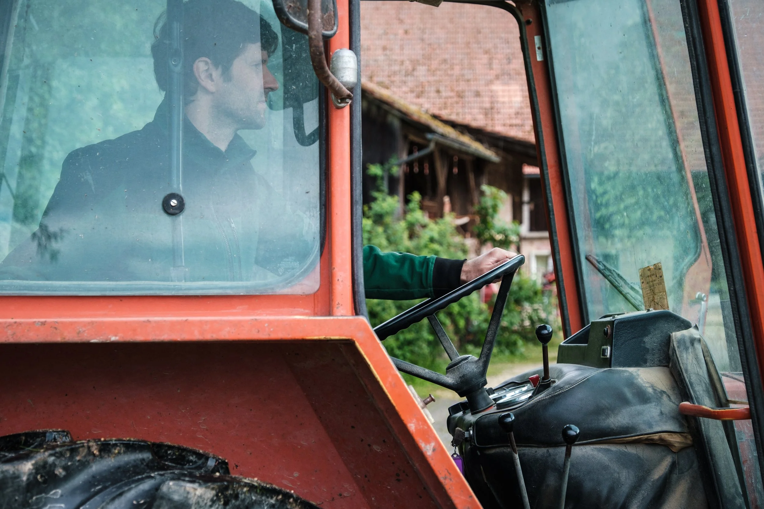 Ein Mann fährt einen roten Traktor, im Blick durch das Fenster. Im Hintergrund sind Häuser und Bäume zu sehen.