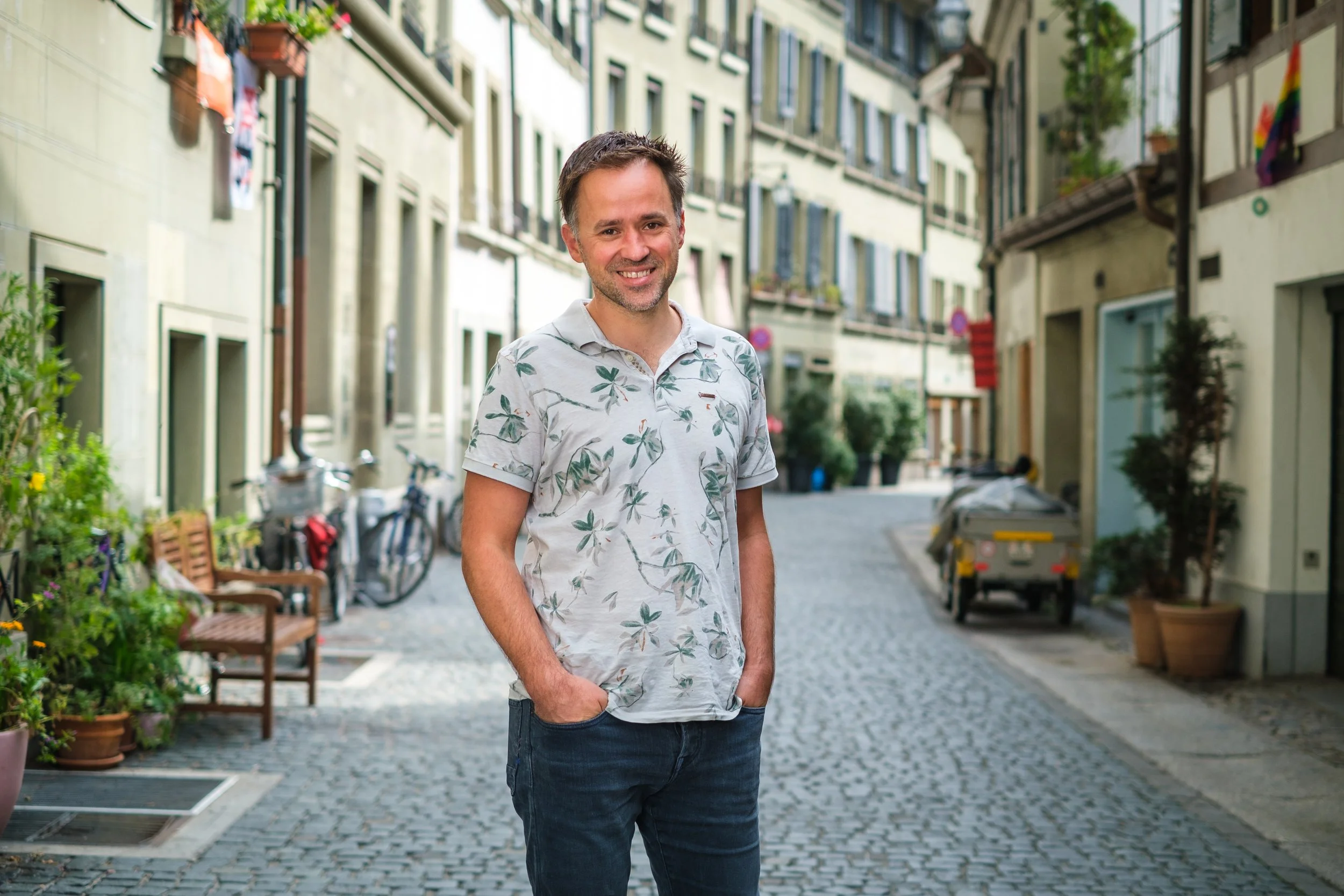 Ein lächelnder Mann steht auf einer gepflasterten Straße in einem europäischen Stadtviertel mit alten Gebäuden, Pflanzen und Fahrrädern im Hintergrund.