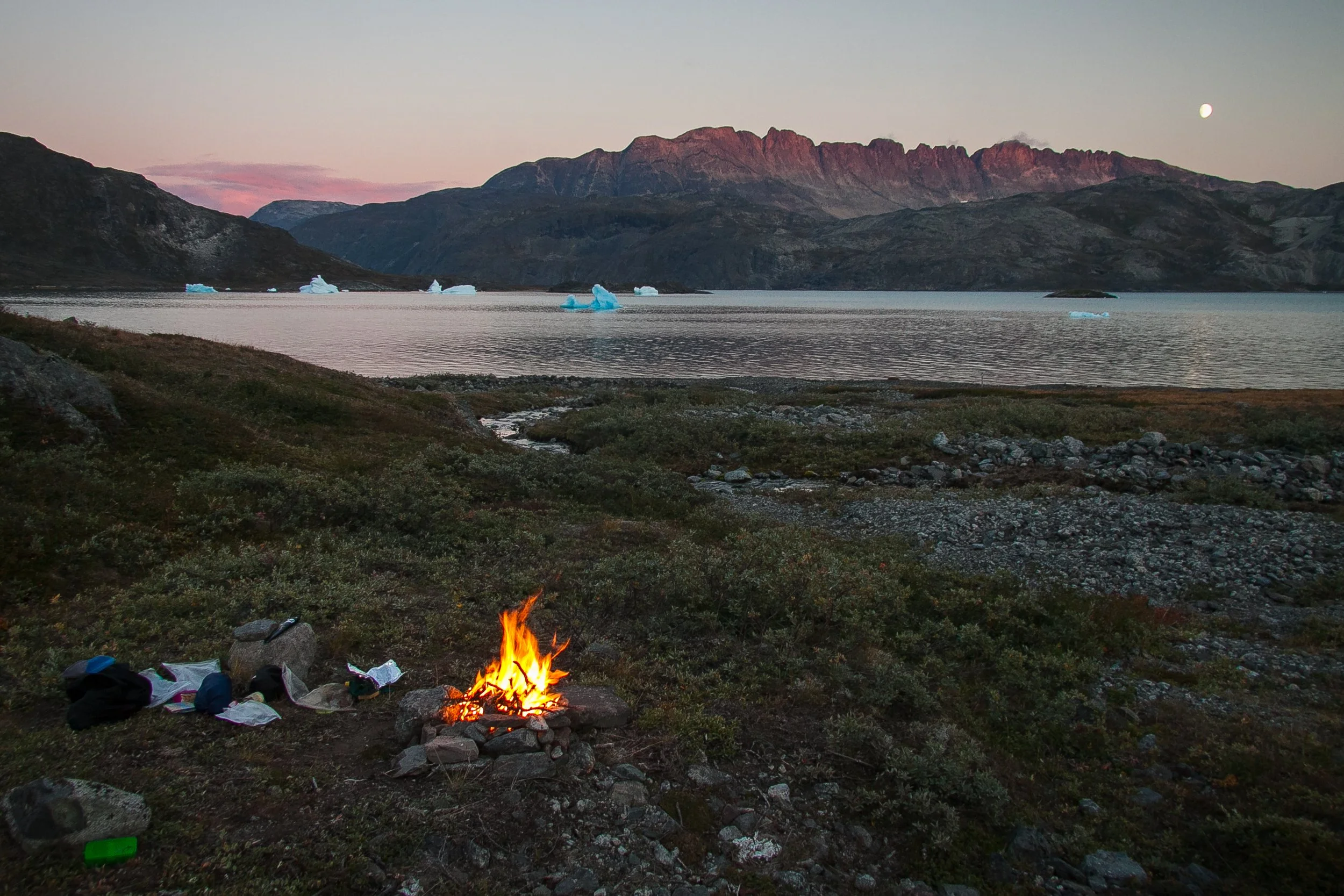 Lagerfeuer im Freien am Ufer eines Sees, umgeben von Bergen bei Sonnenuntergang oder Dämmerung, mit sichtbarem Mond am Himmel.