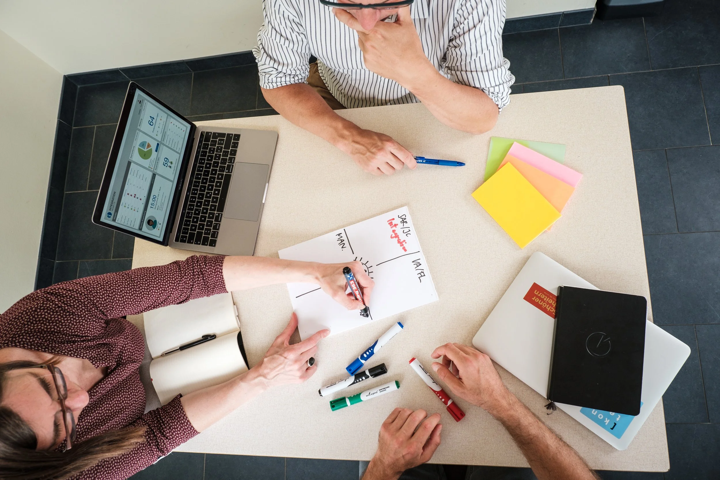Drei Personen arbeiten an einem Tisch mit Laptops, Whiteboard und Marker. Es gibt auch farbige Papierbapiere auf dem Tisch. Die Szene zeigt eine Besprechung oder Teamarbeit.