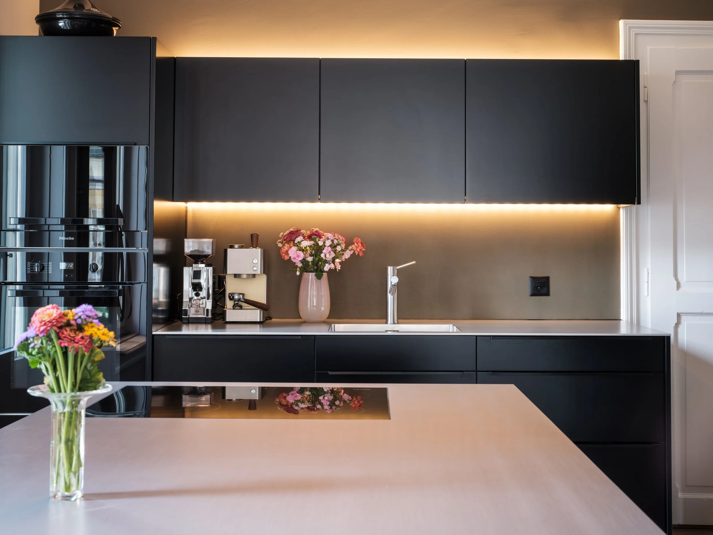 Moderne schwarze Küche mit dunklen Schränken, Kaffeezubehör, Blumenvasen mit Blumen und untergehängter LED-Beleuchtung