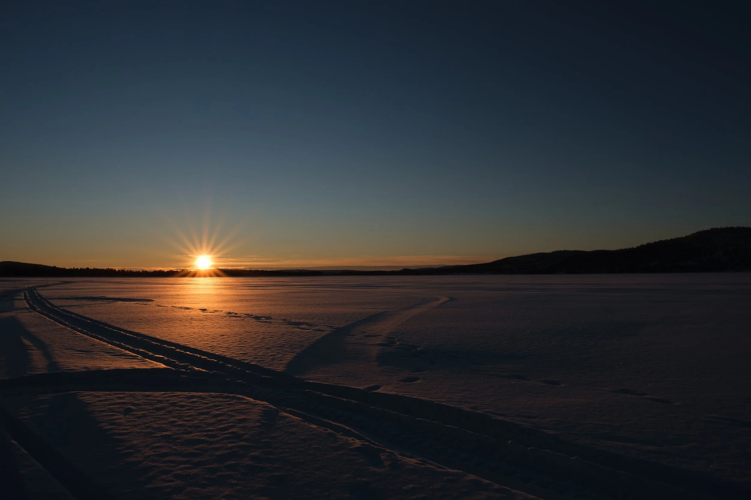 Sonnenuntergang über einem verschneiten, ruhigen See mit Skispuren im Schnee.