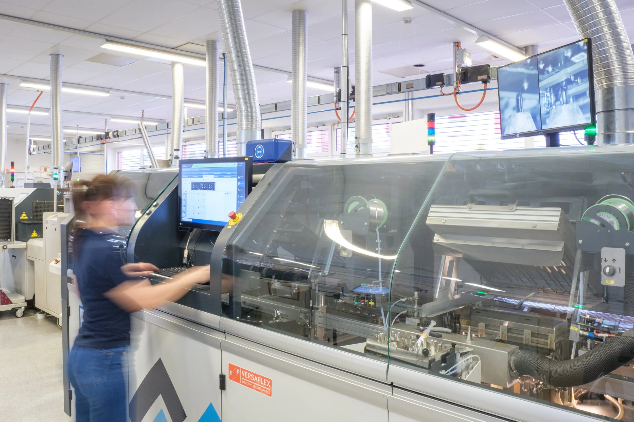 Mitarbeiterin arbeitet an einer automatisierten Maschine in einem Industrieunternehmen, umgeben von technischen Anlagen und Bildschirmen.