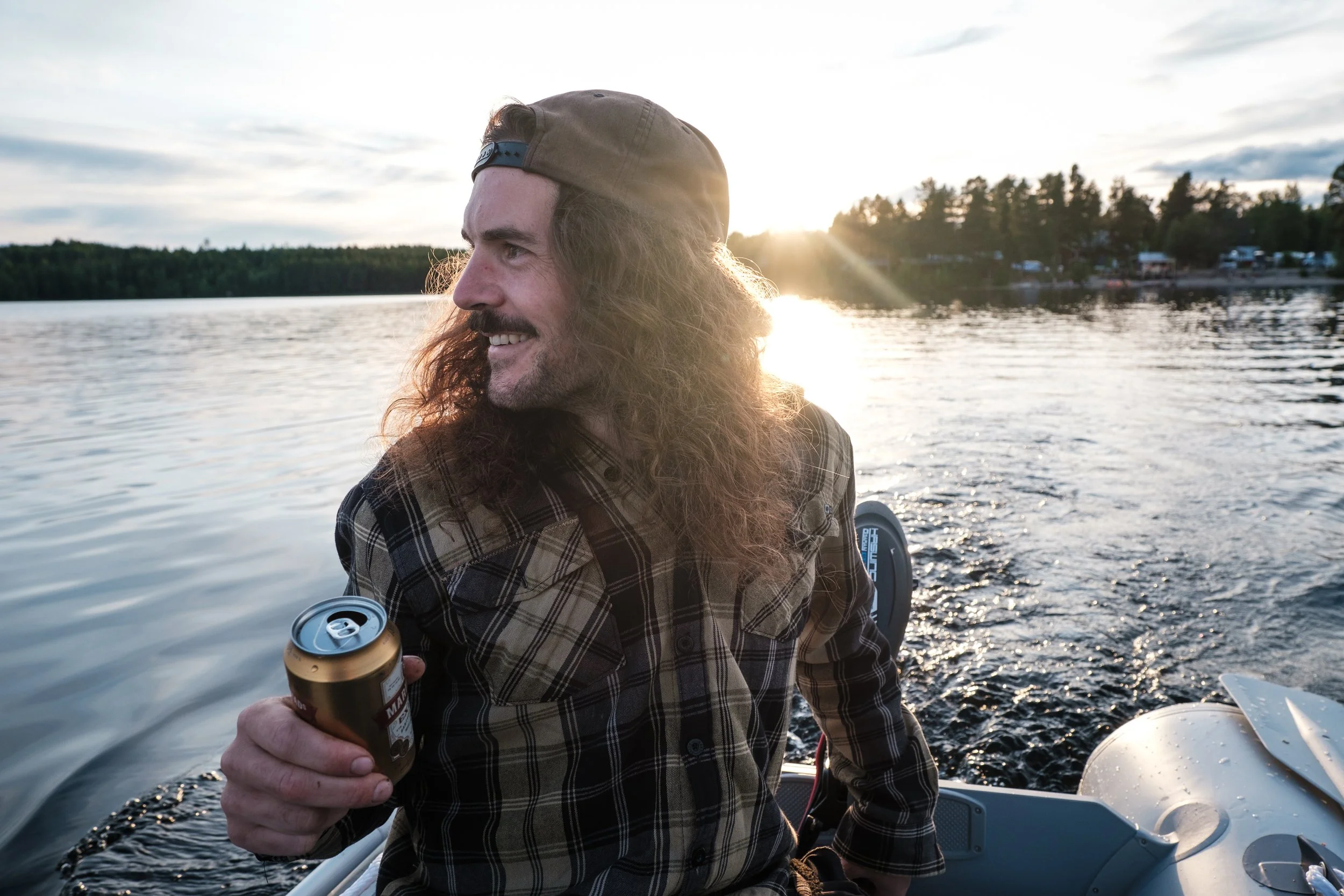Ein Mann mit lang, lockigem Haar und Schnurrbart, trägt ein kariertes Hemd und eine Mütze, sitzt in einem Boot auf einem See bei Sonnenuntergang, hält eine Dose Bier in der Hand und schaut lachend zur Seite.
