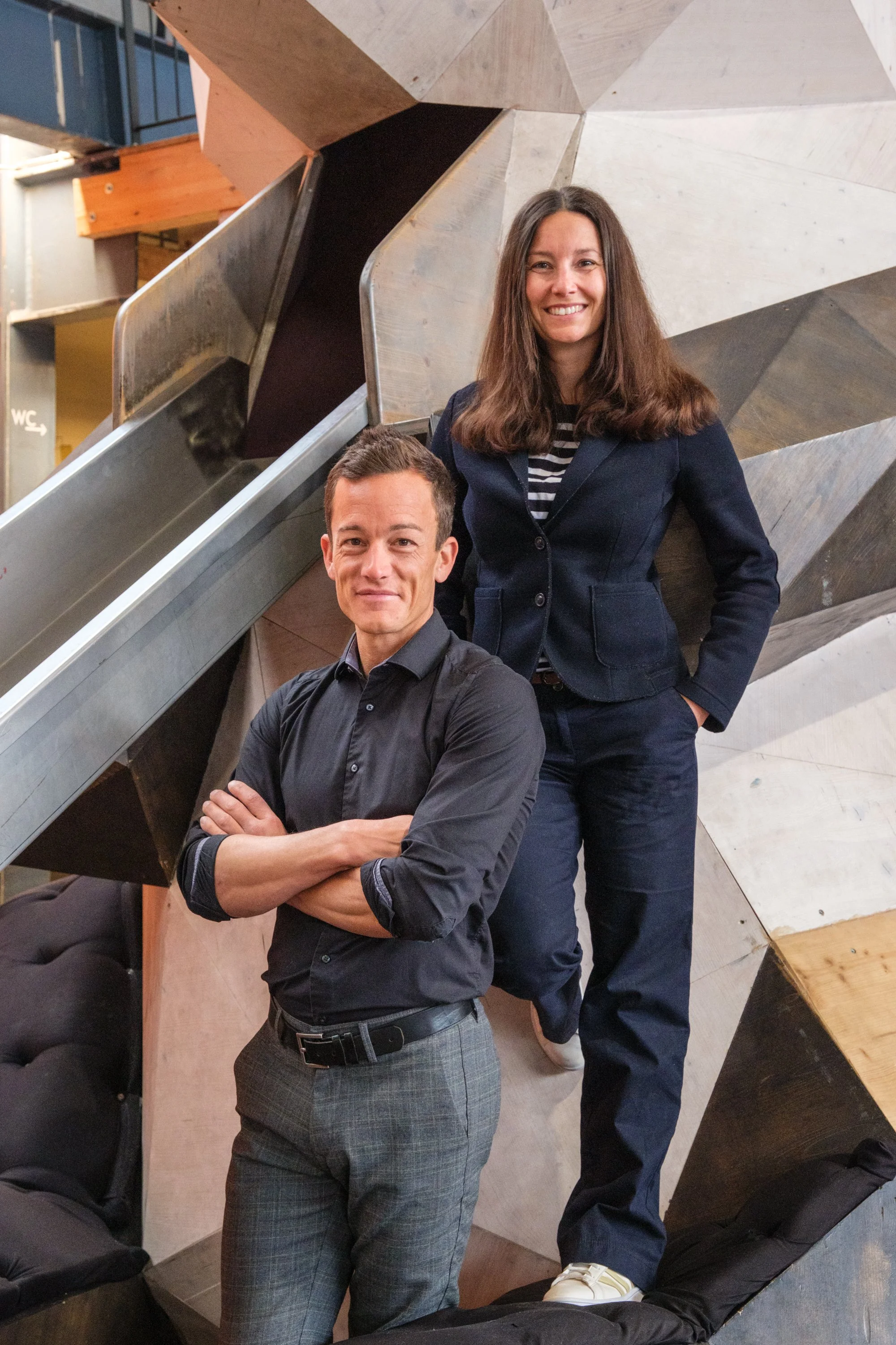 Zwei ehrlich blickende Personen, eine Frau und einen Mann, stehen vor einem modernen, geometrischen Holz- und Metalldesign im Hintergrund.