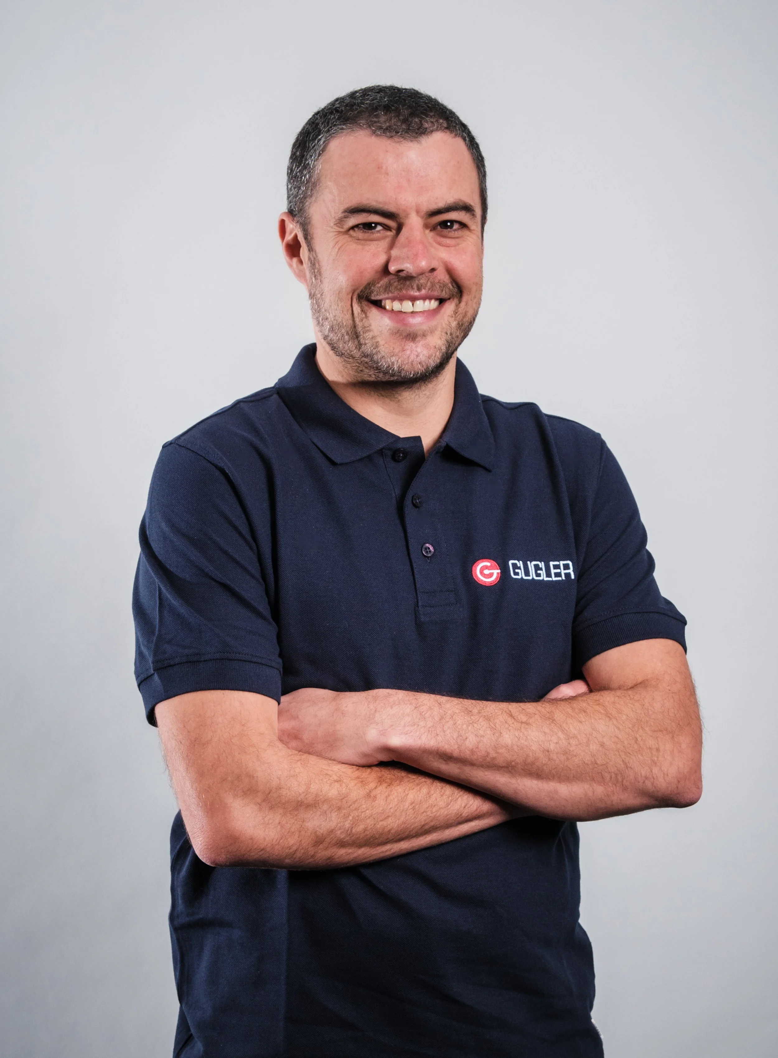 Ein Mann in einem dunkelblauen Polo-Shirt mit dem Logo 'GUGLER', lächelnd vor neutralem Hintergrund, mit verschränkten Armen.
