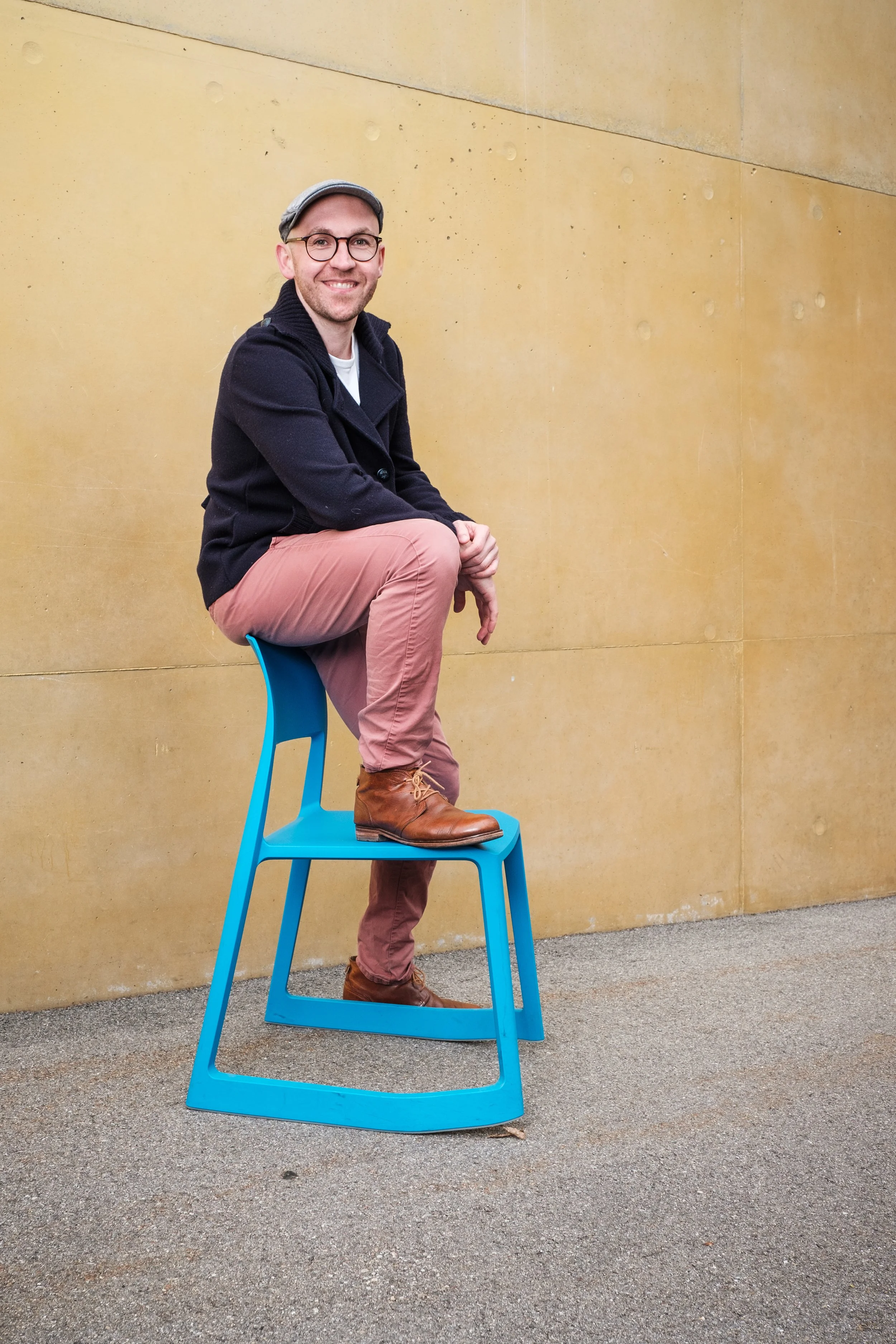 Ein Mann sitzt auf einem blauen Stuhl, der auf einem Straßenboden steht, vor einer gelben Wand, lächelnd in die Kamera schauend.