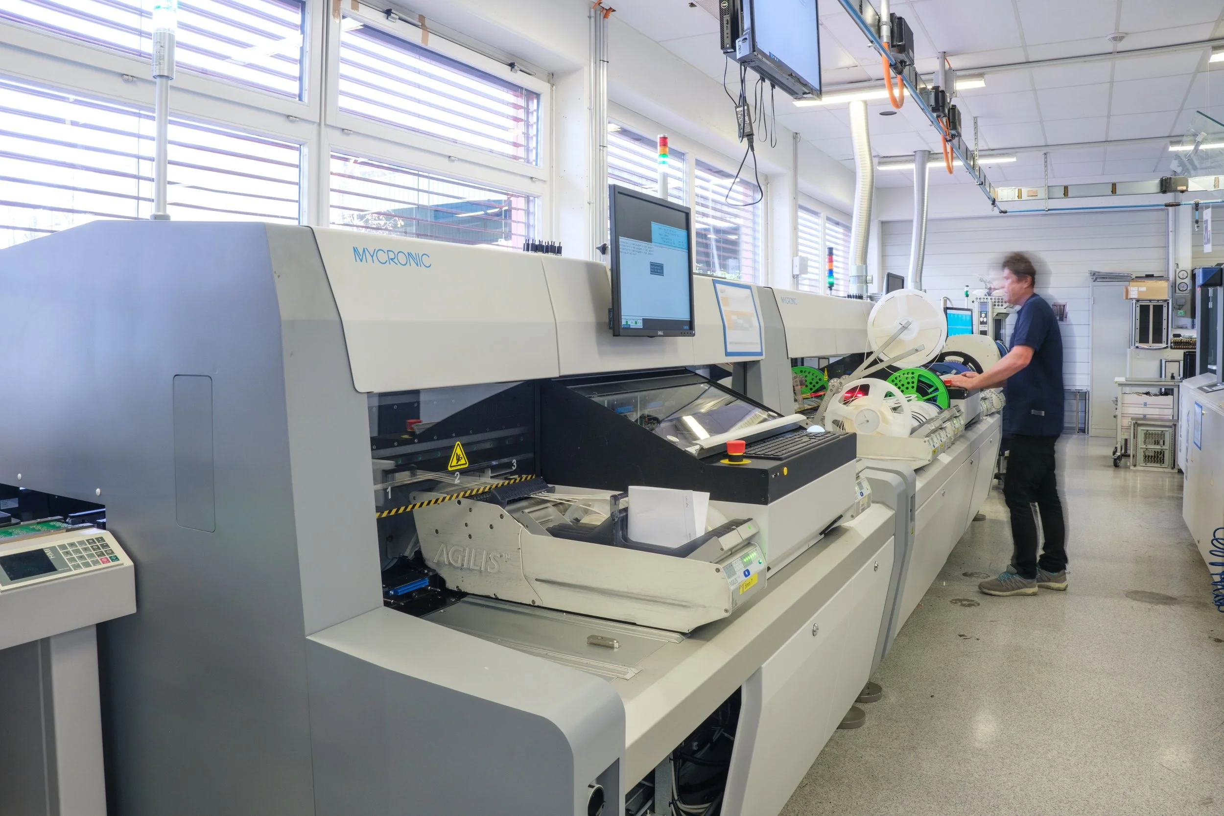 Ein Techniker arbeitet an einer automatisierten Maschine in einem Elektronikfertigungslabor, umgeben von Monitoren und technischen Geräten.