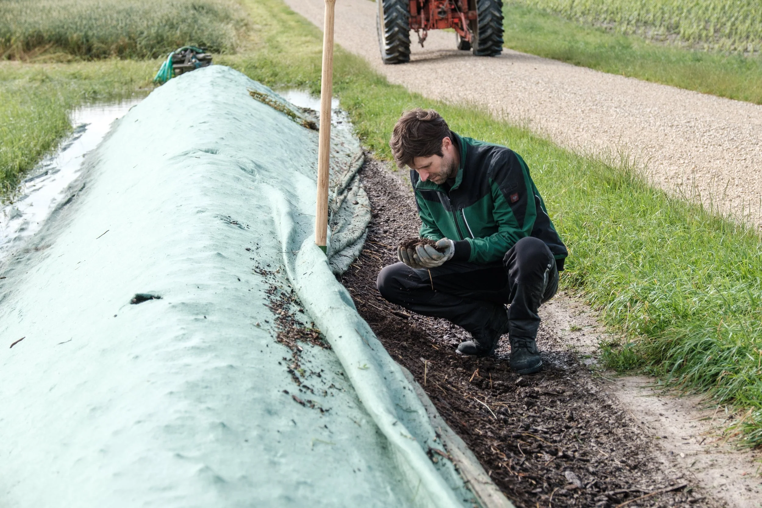Ein Mann kniet neben einer Mulchfolie, die auf einen Mulchberg gelegt wurde, während im Hintergrund ein Traktor auf einem Traktorpfad fährt.