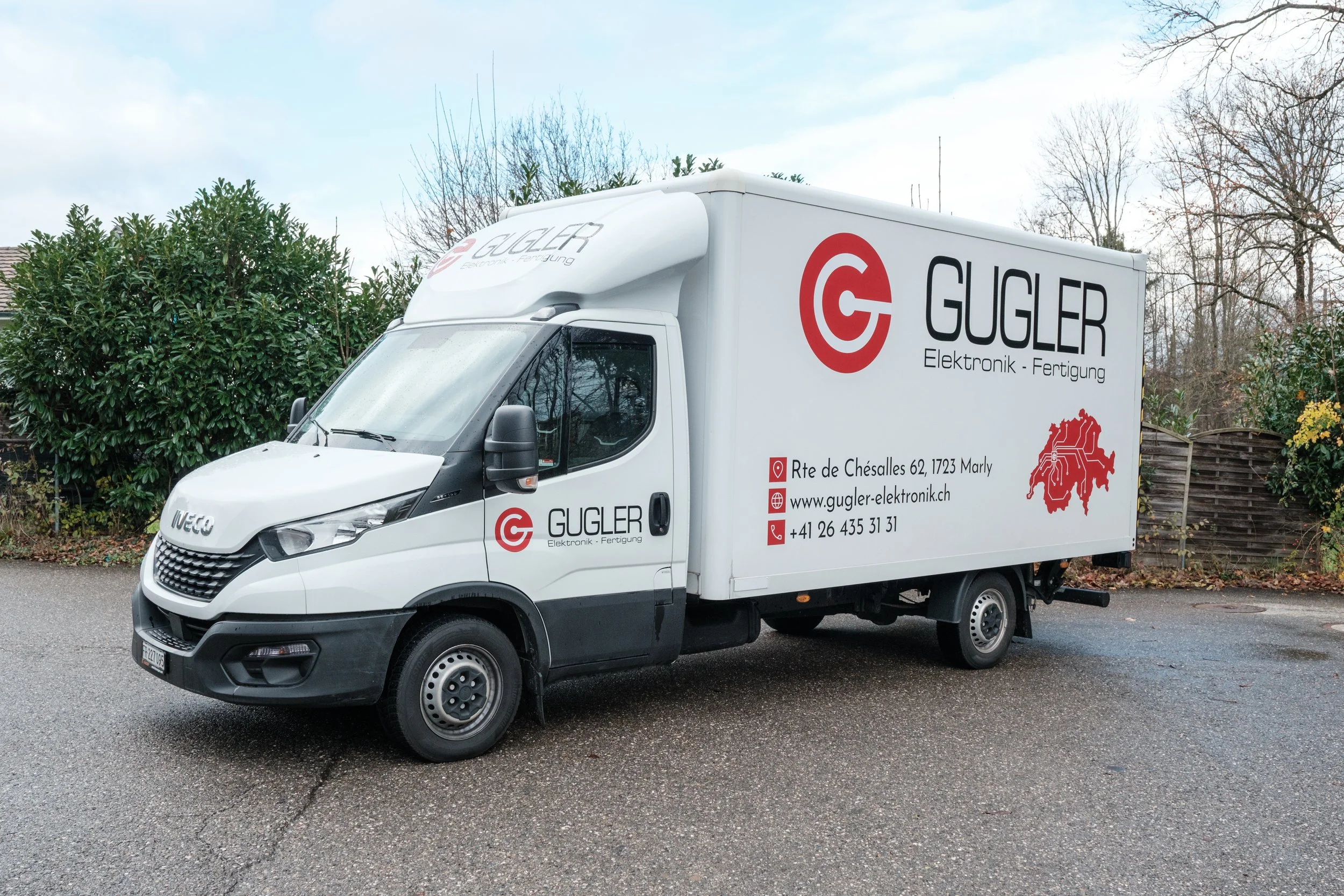 Weißer Lieferwagen mit Firmenlogo und Kontaktinformationen der Firma GUGLER Elektronik - Fertigung, im Freien auf nassem Asphalt bei Bewölktem Himmel, Hintergrund mit Bäumen und Gartenzaun.