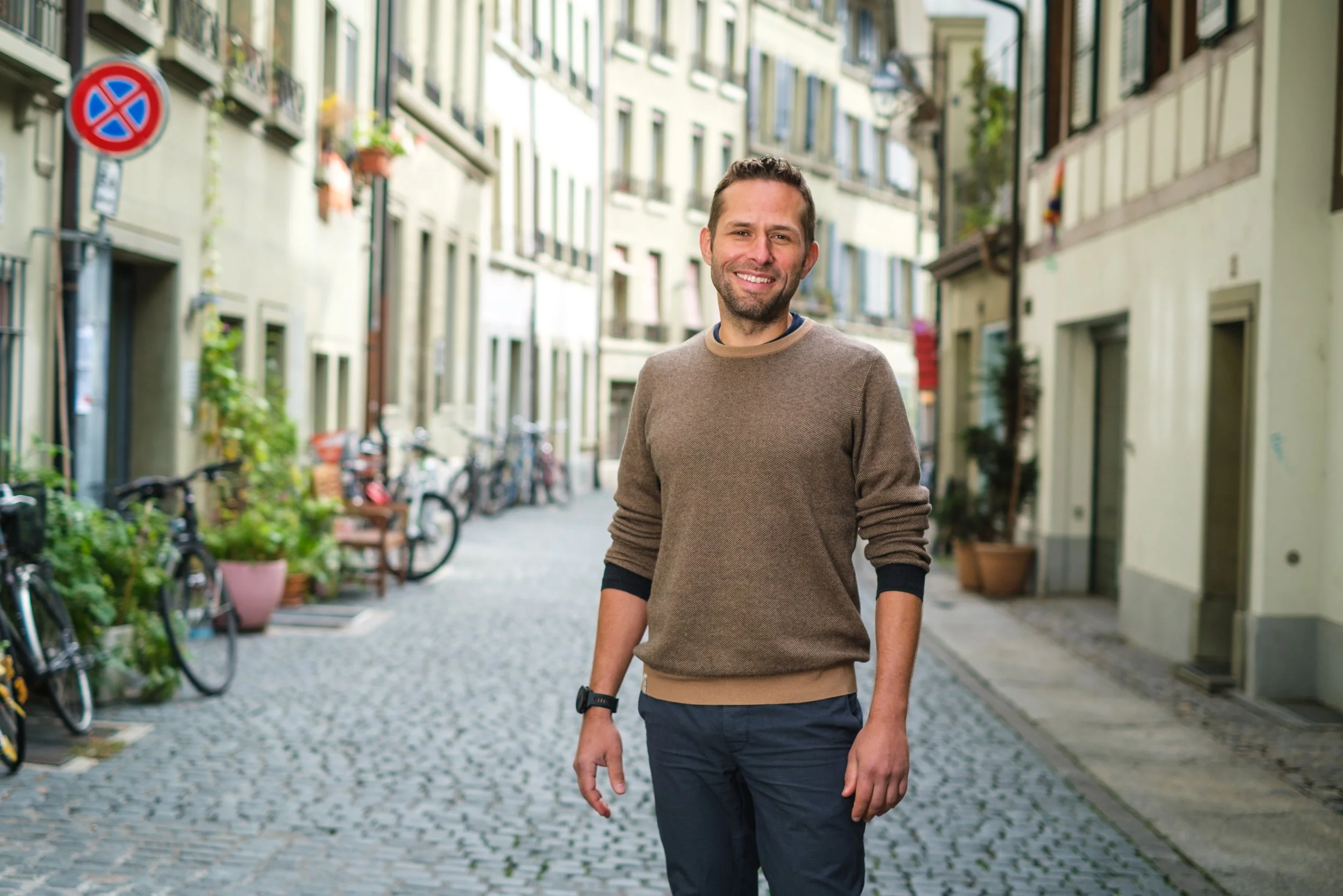 Ein lachender Mann steht auf einer kopfsteingepflasterten Straße in einem Stadtviertel. Im Hintergrund sind Fahrräder und mehrstöckige Häuser mit Fenstern und Pflanzen zu sehen.