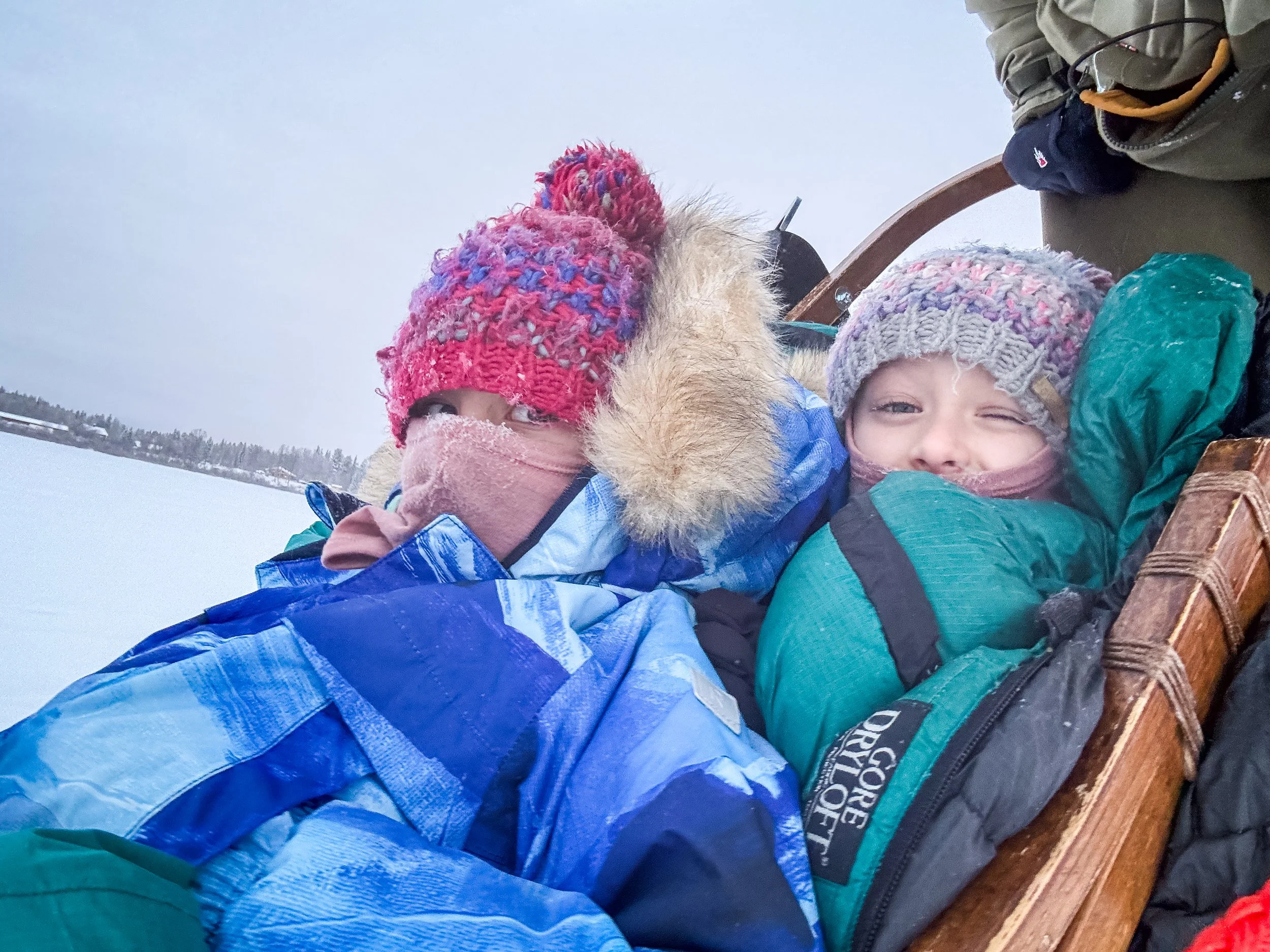 Zwei Kinder in Winterkleidung, sitzen auf einem Schlitten im Schnee, tragen Mützen und Jacken, eine schaut in die Kamera, die andere blickt seitlich, winterliche Landschaft im Hintergrund.