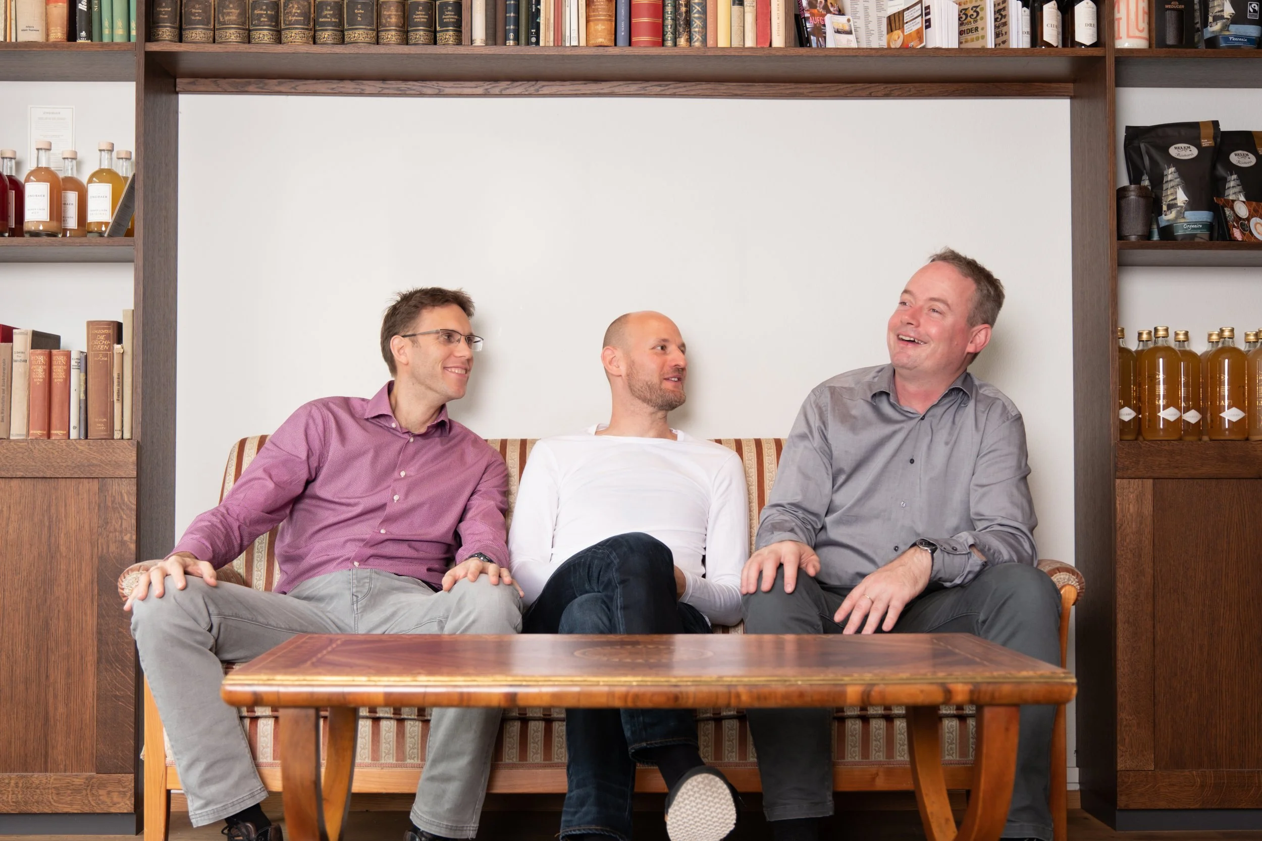 Drei Männer sitzen auf einem Sofa und lachen in einem Raum mit Bücherregalen und einem Holztisch.