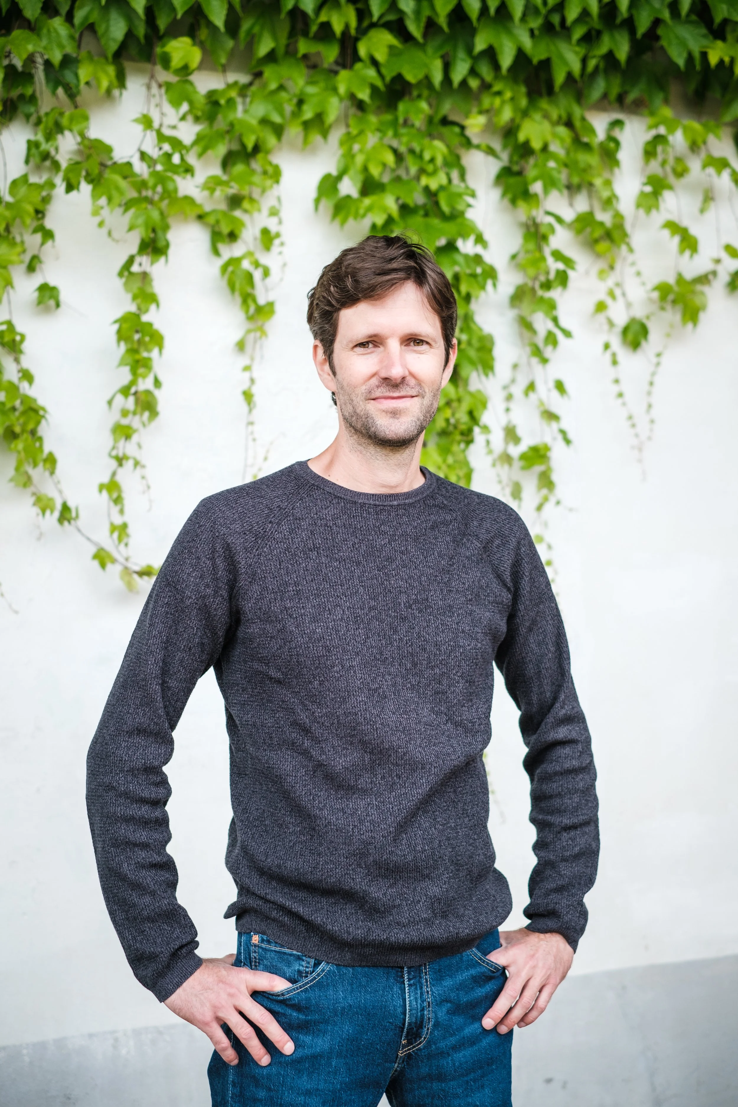 Ein Mann mit braunen Haaren und im grauen Pullover steht vor einer weißen Wand mit grünen Efeuranken.