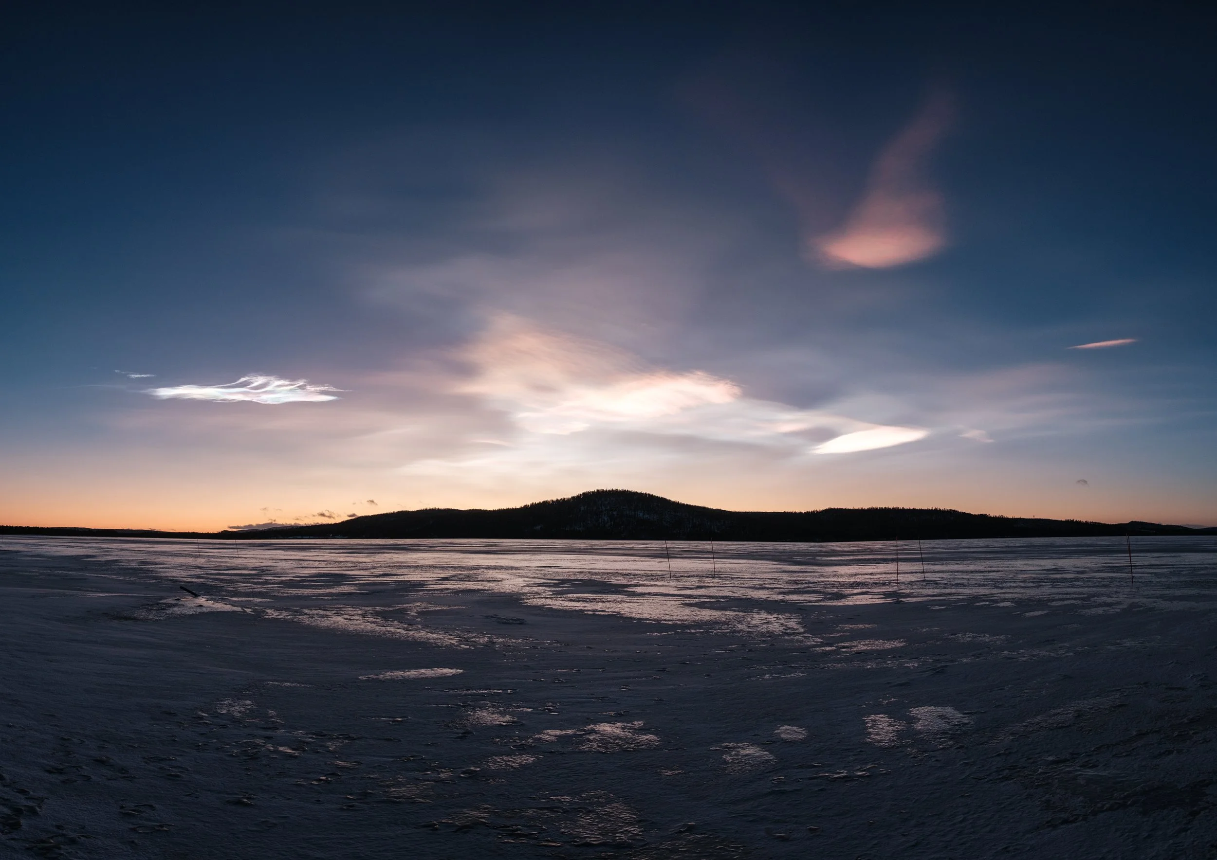 Sonnenuntergang über vereistem See mit dunklem Hügel in der Ferne, gefrorene Oberfläche mit Spuren im Schnee.